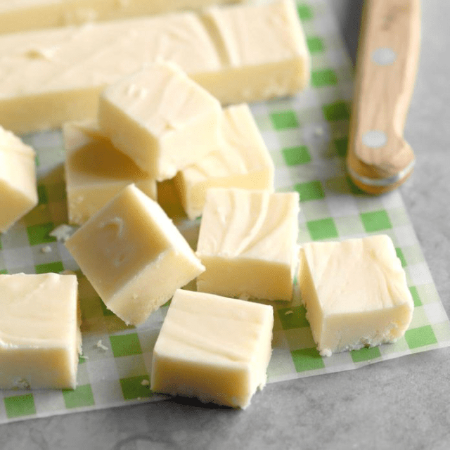 Smooth creamy white chocolate fudge cut into small squares and lying on a green checkered paper.